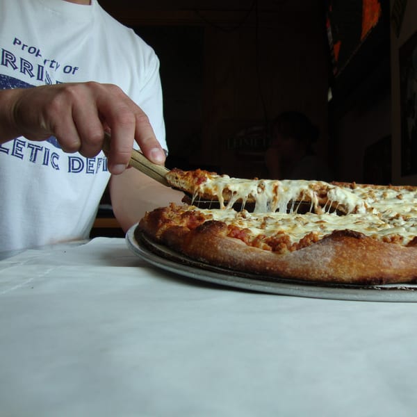 A hand in a white shirt with "Harris" printed on it, serving a cheesy slice of Harris Pizza.