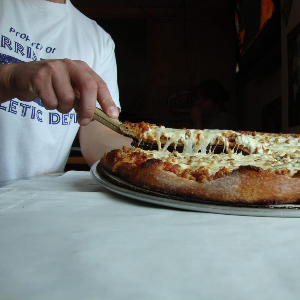 A hand in a white shirt with "Harris" printed on it, serving a cheesy slice of Harris Pizza.