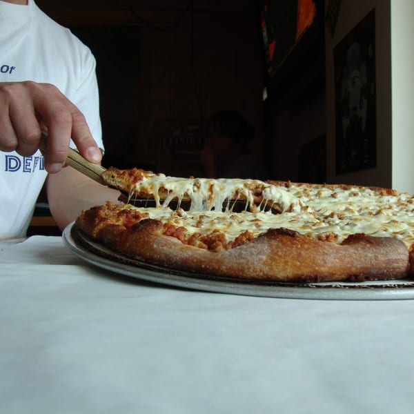 A hand uses a pizza server to lift a cheesy slice of Harris Pizza from a whole pie.