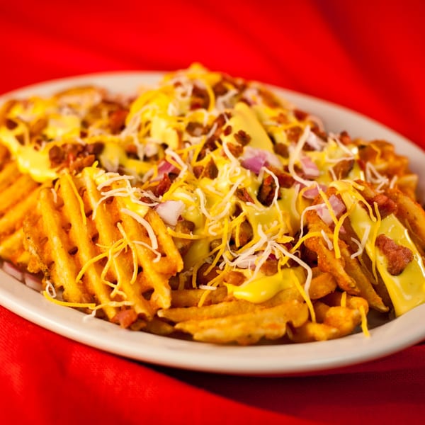 Waffle fries loaded with melted cheese, bacon bits, and red onions on a white plate.