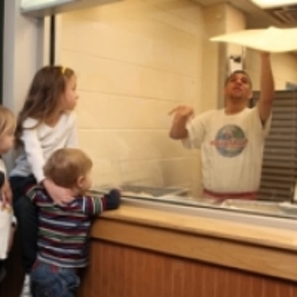 Children watch a Harris Pizza chef toss pizza dough behind a counter.